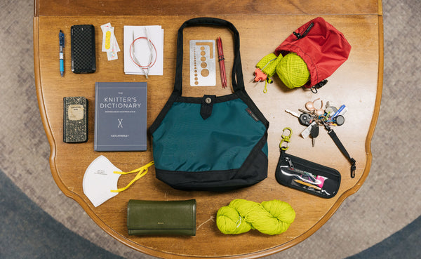 A Little Swift with its various contents fashioned around it: a knitting book, a notepad, a mask, a pair of glasses, a skein of yarn, a knitting tool pouch, a set of keys, cable knitting tools, and a yarn stuff sack with a ball of yarn in it.